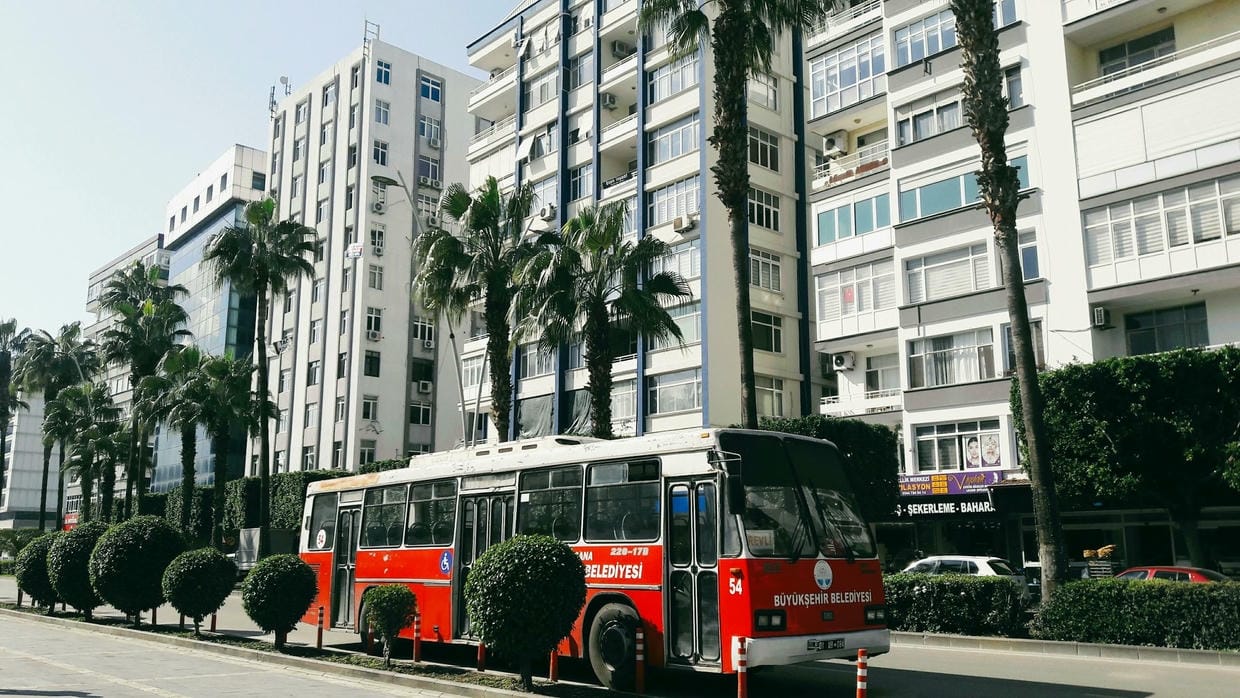 Un autobús urbano en una calle soleada de Adana, Türkiye, con palmeras y edificios urbanos.