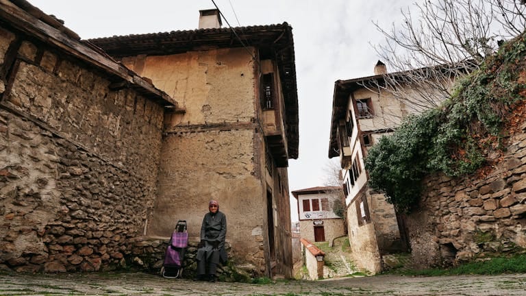 An elderly woman sits on a bench in the historic streets of Safranbolu, Türkiye, showcasing traditional architecture.